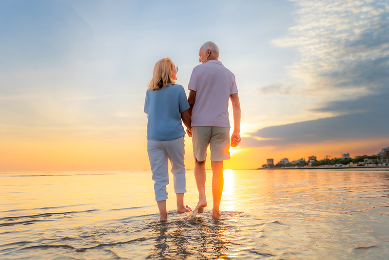 Rear view of senior couple with holding hands walking on beach while vacation during sunset.